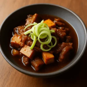 Braised beef tendon with tofu cubes and green onion garnish served in a dark bowl.