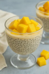 A clear glass filled with mango chia seed pudding and topped with fresh mango cubes on a light blue background.