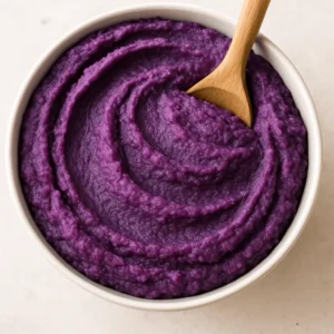 A bowl of creamy purple sweet potato mash with a wooden spoon, featured in a purple sweet potato recipe.