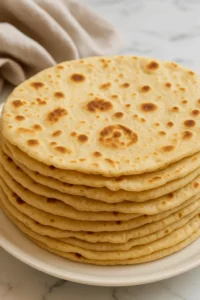 Close-up stack of sourdough discard tortillas on a white plate with soft folds and golden spots