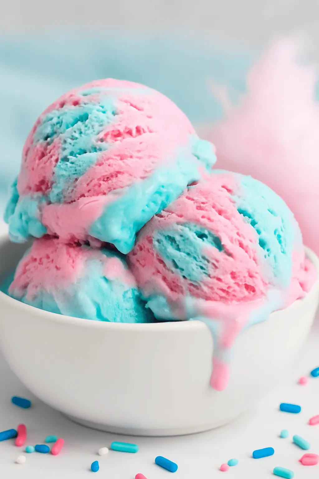 Two scoops of pink and blue cotton candy ice cream in a white bowl with sprinkles scattered around.