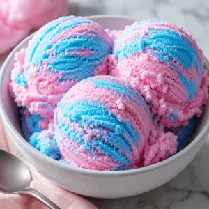 Three scoops of pink and blue cotton candy ice cream in a white bowl with a spoon and soft pink napkin.