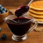 Glass gravy boat filled with thick blueberry syrup, surrounded by fresh blueberries and pancakes — perfect for a homemade blueberry syrup recipe.