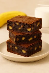Three stacked banana brownies with visible banana chunks and chocolate chips on a plate — a rich banana brownie recipe.