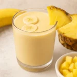 A glass of pineapple banana smoothie topped with banana slices and garnished with a pineapple wedge, surrounded by fresh banana and pineapple pieces.
