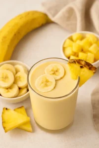 Glass of pineapple banana smoothie garnished with banana slices and a pineapple wedge, surrounded by fresh cut fruit.