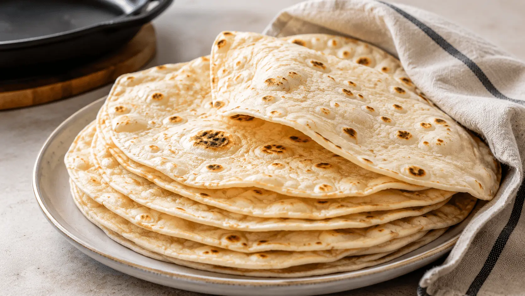 Soft sourdough discard tortillas stacked on a ceramic plate with golden-brown skillet spots and a folded tortilla on top.