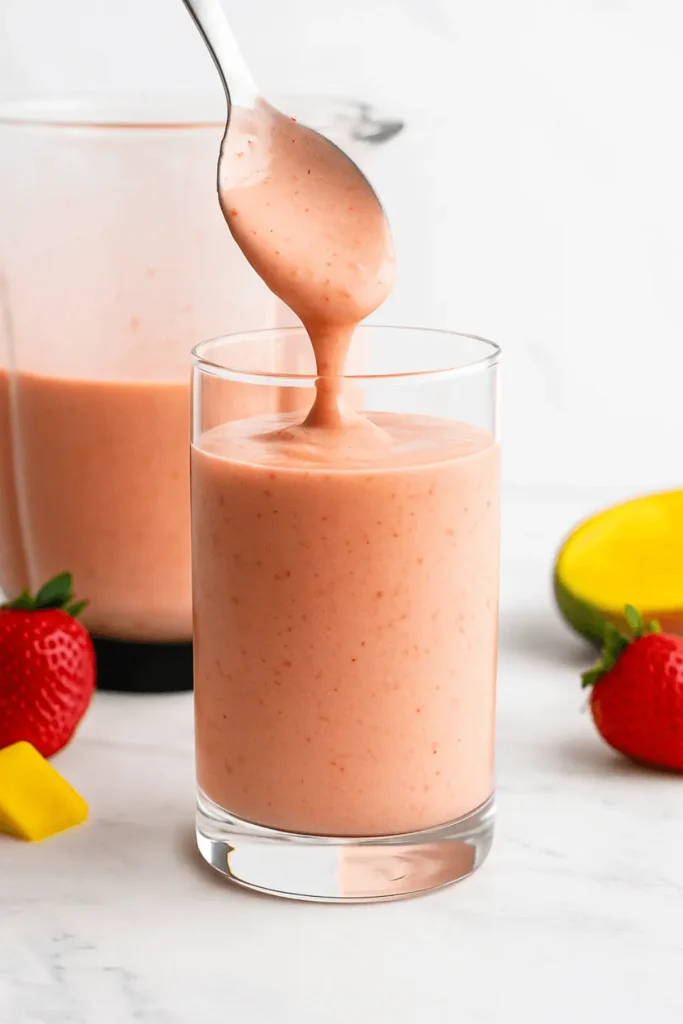 Pouring a thick strawberry mango smoothie from a spoon into a clear glass surrounded by fresh fruit.