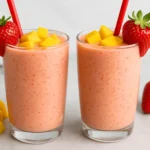 Two glasses of strawberry mango smoothie topped with fresh mango cubes and strawberries on a light background.