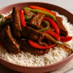A bowl of pepper steak recipe served over fluffy white rice with colorful bell peppers and a glossy brown sauce.