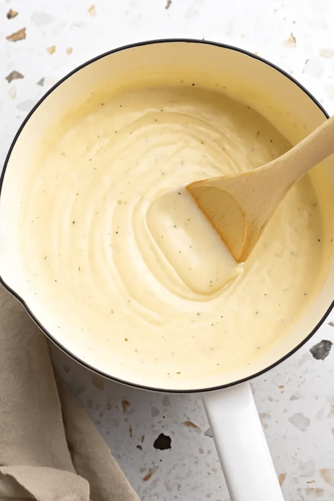 Creamy Alfredo sauce recipe being stirred in a white saucepan with a wooden spoon