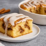 Freshly baked cinnamon roll cake slice drizzled with icing on a white plate.